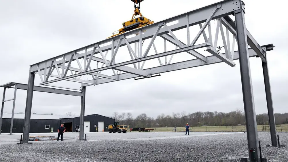 Telehandler with spreader bar lifting a long steel roof truss onto posts of a pole barn under construction