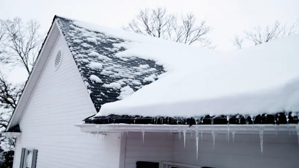 Snow-covered asphalt shingle roof with icicles at the eaves on a two-story house in winter